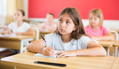 Schoolgirl sitting at desk in classroom during lesson. Her classmates sitting around.