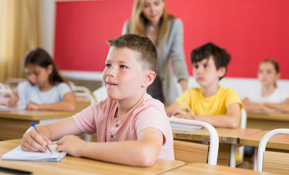 Boy and his classmates sitting at desks in classroom. Their teacher standing in background.