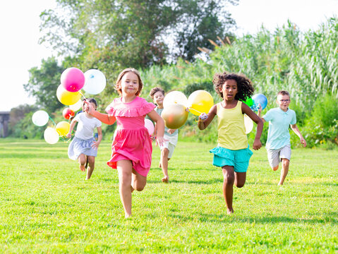 Portrait of cheerful preteen boys and girls with colorful toy balloons in hands running on green lawn in city park on sunny summer day
