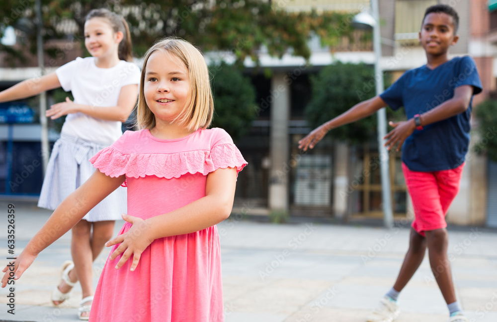 Wall mural Positive cute girl dancing modern choreography with group of tweenagers on city street on summer day..