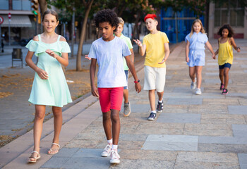 Cute preteen girl talking friendly with her african american friend while walking together along city street on summer day..