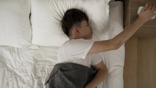 view from above sleepy asian man unable to wake up is sleeping in after stopping the ringing electric alarm clock in the morning on bed in the bedroom at home