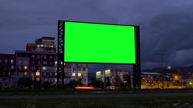 Timelapse: blank green billboard, large display, fast moving car traffic in evening time with warm night illumination, gets dark. Green screen, time lapse, template, mock up and copy space concept