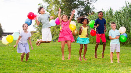 Barefoot children jumping on field with balloons in hands and smiling.