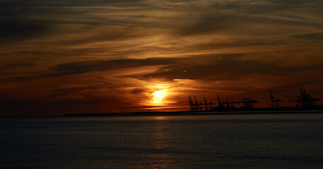 Fototapeta na wymiar Distant sunset with orange sky and clouds over the sea, with Le Havre port cranes in the background
