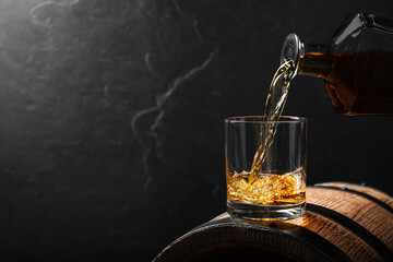 Pouring whiskey into glass from bottle on wooden barrel against grey background, space for text