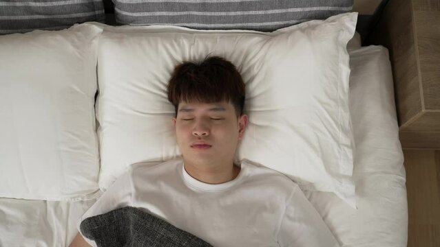 top angle shot with zoom in restless asian boy awaking to bad dream is patting his chest and staring upward at camera while breathing heavily in bed at home