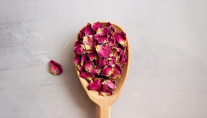 Dried rose petals in a wooden spoon, top view. Light background, copy space. Pink colored organic herb used for perfumes, cosmetics, teas and baths.