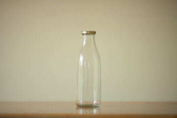 One empty glass bottle for milk standing on the wooden table over light beige background. Day light. Copy-space. Indoor shot