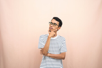 Asian man in glasses wearing casual striped t-shirt, gesturing get ideas, looking for ideas, thinking. Isolated beige background.