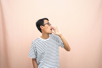 Asian man in glasses wearing casual striped shirt, shouting pose with hand near mouth, giving information. Isolated beige background.