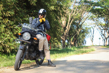 Full body size photo of man sitting on a motorcycle, wearing sunglasses and a black jacket, fastening his helmet with a long road in the background.