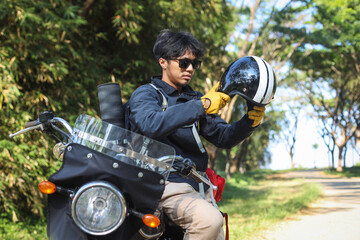 Man sitting on a motorcycle, wearing sunglasses and a black jacket, holding his helmet with a road in the background.