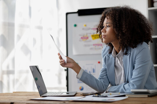 American woman sitting working in a startup company, american businesswoman working in the office, she is looking at financial documents and sales data to summarize before meeting with executives.