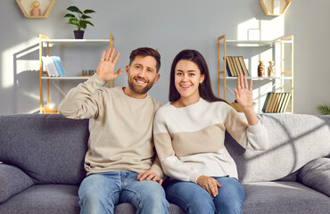 Portrait of a happy smiling couple waving hands and smiling to camera sitting on sofa at home. Man...