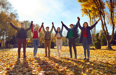 Happy friends standing together raising their hands in park. Group of young people wearing warm...