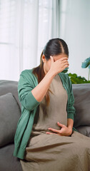 Asian pregnant woman having nausea and headache while relaxing on sofa at home.