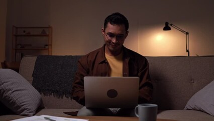 lucky cheerful Asian guy sitting on sofa using laptop and fists raised after receiving good news in the Living Room.