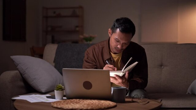 Asian man freelancer study or learn in his laptop computer at  with notebook sitting on sofa at home Online e-learning at night room.