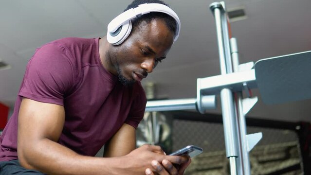 Tired black guy bodybuilder sitting at gym, listening to music, using mobile phone and wireless headset. African american young man with headphones holding smartphone