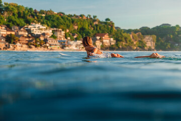 Surfer woman rowing on surfboard in ocean with coastline landscape on background.