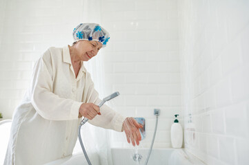 Elderly woman in bathrobe filling bathtub with water - obrazy, fototapety, plakaty