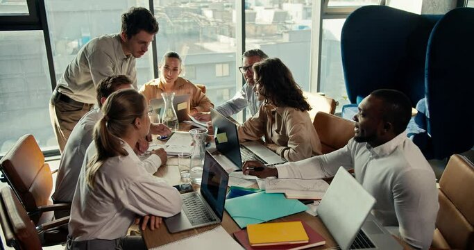 A group of office workers sit at a table and show the manager guy in a white shirt their projects on paper. Team work at a table in an office with panoramic windows
