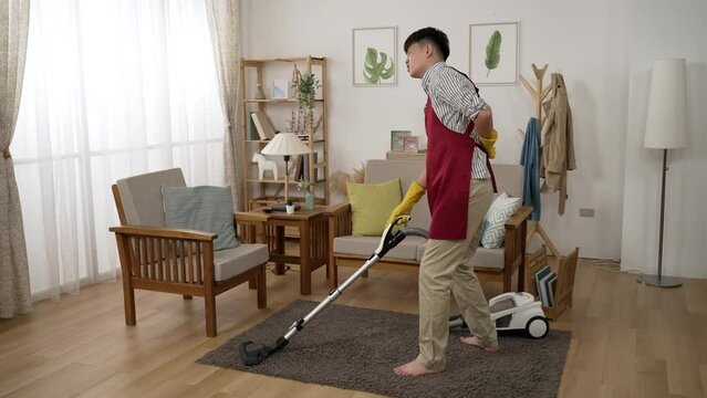 full length with side view asian man housekeeper in gloves and apron sprains his lower back and moans painfully while he is vacuuming floor in the living room