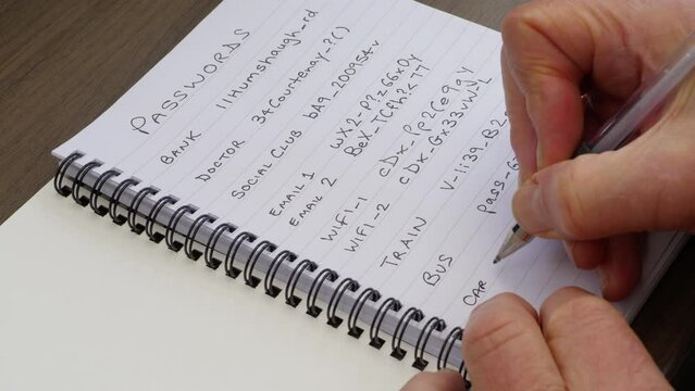 Closeup of a man's hands opening a lined notebook, then using a pen to write another online password, to add to the variety of logins already listed above.