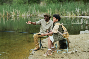 African American son fishing together with his dad while they sitting near the lake