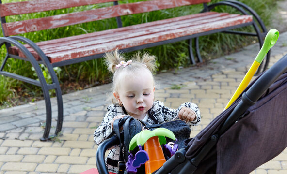 Baby pushing a stroller on a walk in summer park. Adorable little girl near pushchair and waiting for mom