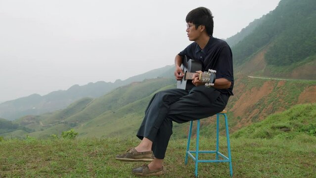 Talented asian musician man playing the guitar sitting on a stool in middle of green mountains