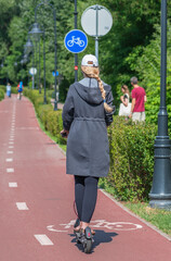 Girl with blonde hair riding an electric scooter on the bike road with blue road sign or signal of bicycle lane among green trees and hedges, spring summer nature, vertical 