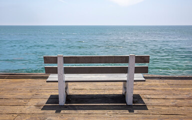 Fototapeta na wymiar Empty wooden park bench on waterfront wood platform. Sunny day, calm sea, blue sky background. USA