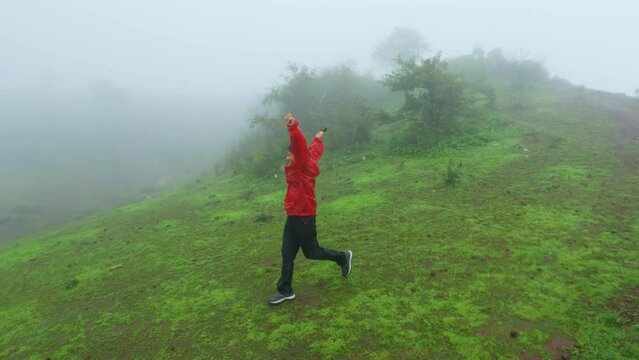 Teenager girl wearing red rain jacket running on the lush green hill at Saputara Hill Station, Gujarat, India. Mountain covered by green grass on both side during monsoon season.