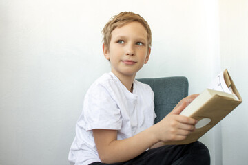 Cute boy reading a book while sitting on a chair and looking away