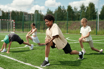 Group of schoolchildren doing physical exercise on green grass of football field or stadium while repeating it after African American schoolboy
