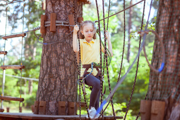 Naklejka na ściany i meble Child in a forest adventure park made of ropes. Children's outdoor climbing entertainment center. Playground for children and sports with a cableway. Sports suspended rope trainer.