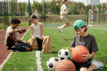 Cute schoolboy looking at basketball ball in his hands while choosing item for playing sports game against intercultural classmates or friends