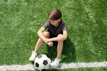 Above view of tired schoolboy with soccer ball between feet sitting on green lawn at stadium and...