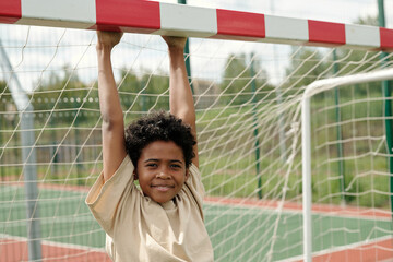 Cute smiling schoolboy in t-shirt hanging on goal gate and looking at camera while enjoying leisure...
