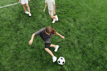 Above angle of active schoolboy kicking soccer ball while moving along green field and playing...