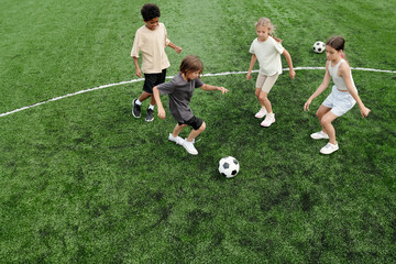 Above view of four active kids in sportswear running along green football field while playing soccer together at stadium on weekend or at leisure