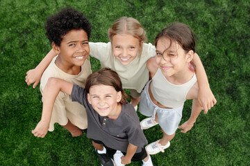 Fototapeta premium Above angle of several happy intercultural children in activewear standing against green lawn or football field and looking at camera with smiles