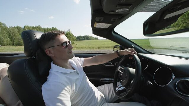 Smiling man driving car by grassy landscape with sky in background. Handheld shot of young male enjoying road trip during summer. He is wearing sunglasses during sunny day.