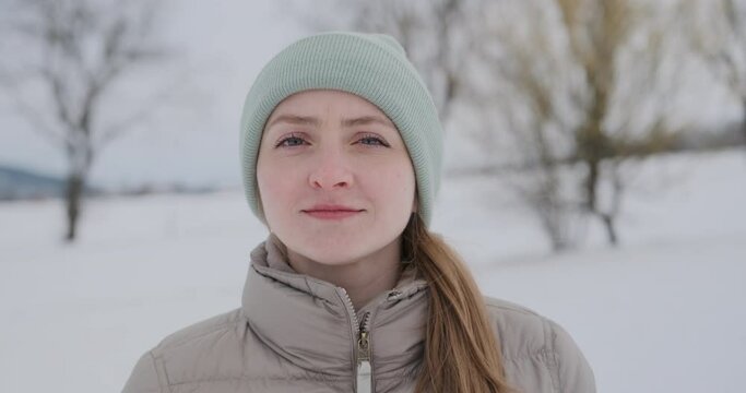 Young woman with fair skin in hat and warm jacket against the backdrop of winter landscapes looks at the camera and smiles.