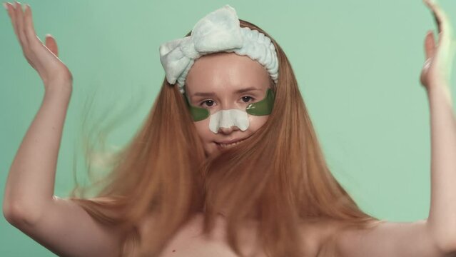 A woman with hydrogel patches under her eyes and a patch for blackheads on her nose on a green screen. A woman demonstrates healthy hair tied with hair band. Advertising area. Slow motion.