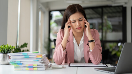 A stressed Asian businesswoman sits at her desk with an unhappy face, suffering from headache