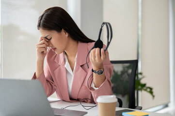 A stressed Asian female call centre agent sits at her desk, feeling tired and overwhelmed.