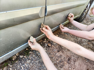 Hands of children with a stone scratching or drawing stripe or pattern on the side of the car. The...